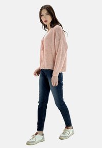 Helles rosa gestricktes Cardigan mit lockerer Passform, kombiniert mit dunkelblauen Jeans und weißen Sneakers mit braunen Akzenten, vor einem schlichten Hintergrund.