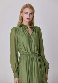 Femme blonde portant une robe à manches longues de couleur vert olive clair avec des détails floraux au col et une ceinture nouée à la taille, sur un fond uni.