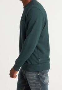 Mörkgrön sweatshirt i texturerad tyg med rund hals och ribbad mudd. Har en liten logotyp patch på bröstet och sidoflikar.