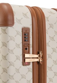 Harde koffer in roomkleur met een subtiel patroon, bruine accenten, dubbele ritsen en een cijferslot. Beschikt over gouden hardware-details.