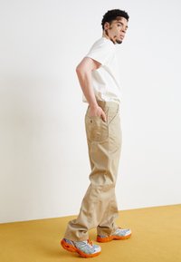 Beige pants with a straight cut, featuring a back pocket with a logo, paired with colorful, patterned sneakers on a yellow surface.