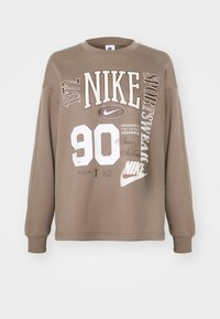 Brūns kreklu džemperis ar baltu grafisko tekstu, tostarp "NIKE", "90" un dažādiem logotipiem. Materiāls izskatās mīksts ar brīvu piegriezumu.