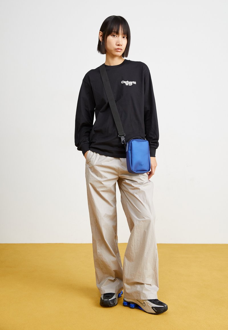 Sac en nylon bleu porté en bandoulière avec une fermeture éclair, associé à un tee-shirt noir à manches longues et un pantalon ample de couleur claire, ainsi qu'à des baskets.
