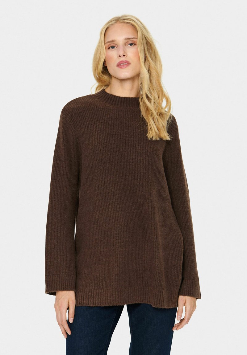 Brūns adīts džemperis ar brīvu piegriezumu, augstu apkakli, ribotu tekstūru un garām piedurknēm, saskaņots ar tumši ziliem džinsiem.