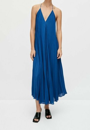 Kvinde iført en flydende, plisseret, ærmeløs blå midikjole med tynde stropper, kombineret med sorte åbne slip-on sko.