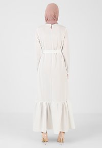 Long, white dress with a flared hem, belted waist, long sleeves, and back keyhole detail. Smooth fabric with subtle texture, paired with nude heels.