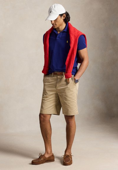 Mann trägt weiße Kappe, blaues Polohemd, roten Pullover über die Schultern gelegt, beige Shorts, braunen Gürtel und braune Bootsschuhe.