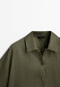 Chemise en lin vert olive avec un col classique et deux boutons visibles sur fond blanc.