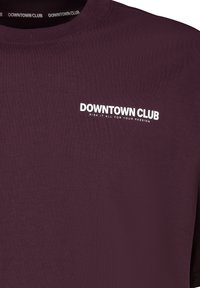 Bordeauxfarbiges Langarmshirt mit Rundhals; das "DOWNTOWN CLUB"-Logo in Weiß mit gut sichtbarem Slogan. Glatte Textur.