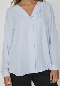 Blouse bleu clair à manches longues avec un décolleté en V, texture lisse et coupe décontractée, dotée d'un ourlet arrondi et de poignets boutonnés.