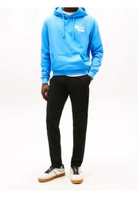 Modrá mikina s klokaní kapsou a kapucí na šňůrku, spárovaná s černými kalhotami a bílými teniskami s černými a hnědožlutými akcenty.