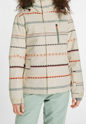 Vrouw draagt een beige winterjas met patroon en een borstzak met rits, en lichtgroene broek, handen zichtbaar langs de zijkanten.