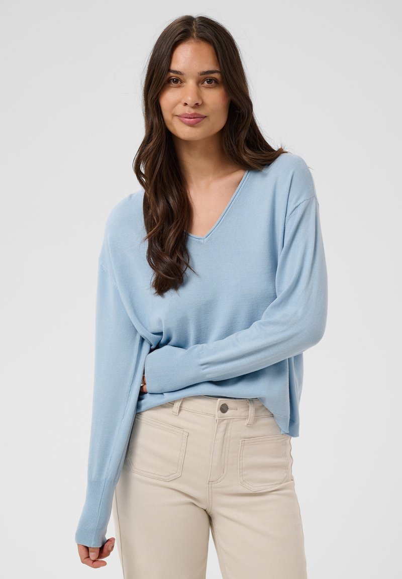 Jeune femme aux longs cheveux bruns portant un pull bleu clair à col en V et un pantalon beige, debout devant un fond clair uni.