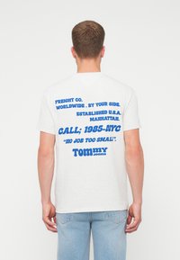 Muž v bielom tričku s modrým textom o Freight Co., Manhattan a Tommy Jeans na chrbte, doplnený svetlomodrými džínsami.