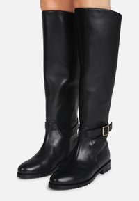 Bottes en cuir noires montantes avec fermeture éclair sur le côté et sangle ajustable, portées sur des jambes nues.