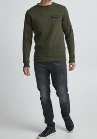 Felpa verde oliva con collo tondo e toppa logo sul davanti, abbinata a jeans grigio scuro e sneakers nere alte. Texture del tessuto liscia.