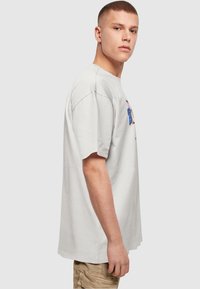 Ung mand med kort hår iført en hvid oversized T-shirt og beige cargo-bukser, set sidelæns mod en ensfarvet baggrund.