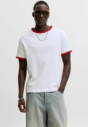 Giovane uomo con occhiali da sole scuri, maglietta bianca con bordo rosso, catena d'argento e jeans azzurri con una mano in tasca.