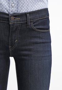Primer plano de unos pantalones de mezclilla azul oscuro combinados con una camisa de patrón azul claro metida por dentro, mostrando los detalles de costura y botones.