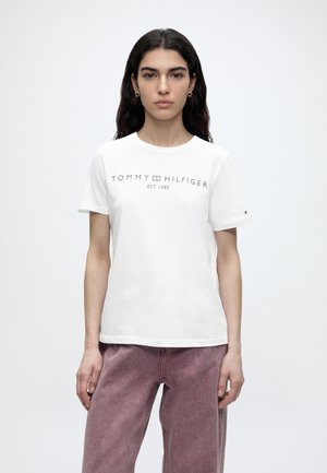 Γυναίκα με μακριά σκούρα μαλλιά, που φοράει λευκό T-shirt Tommy Hilfiger και μωβ παντελόνια με ψηλή μέση, στέκεται μπροστά σε απλό λευκό φόντο.