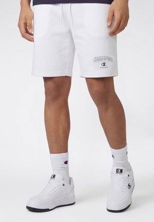 Mężczyzna w białych spodenkach Champion, białych skarpetkach Champion i białych butach sportowych Champion, stojący na jednolitym tle.