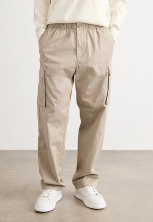 Beige cargo pants with an elastic waistband, two side pockets, and two cargo pockets on the thighs; paired with white sneakers.