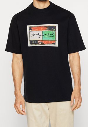 Homme portant un t-shirt noir avec un graphisme rectangulaire multicolore et le texte « Tommy Hilfiger » sur la poitrine.