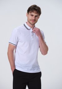 Hvid polo-shirt lavet af bomuldsblanding, med en krave med sorte striber og tre knapper ved halsen. Korte ærmer, afslappet pasform.
