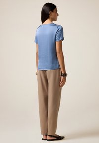 Donna in piedi di profilo che indossa una maglietta blu a maniche corte, pantaloni beige, scarpe flat nere con cinturino alla caviglia e braccialetti spessi su entrambi i polsi.