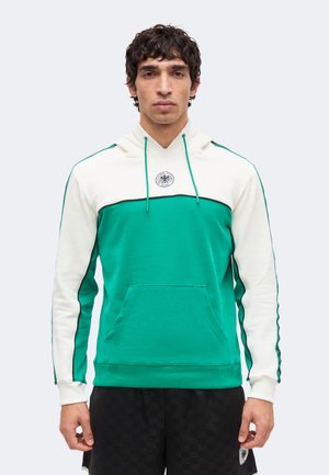 Mies pukeutunut vihreään ja valkoiseen huppariin, jossa edessä tasku, sekä mustiin shortseihin, joissa vasemmassa lahkeessa pieni tunnus.