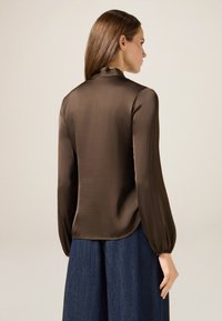 Blusa in raso marrone con maniche lunghe a sbuffo e colletto alto; presenta una texture liscia e un orlo arrotondato, abbinata a una gonna in denim blu.