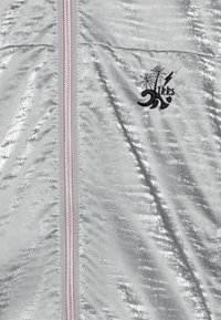 Srebrna kurtka na suwak o błyszczącym, fakturowanym wykończeniu, z różowymi akcentami zamka błyskawicznego oraz czarnym, haftowanym logo z motywem palmy i fali.