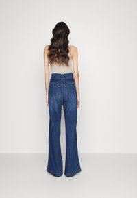 Jeans de mezclilla oscuros de pierna ancha con cintura alta, costura visible y dos bolsillos traseros; combinados con una blusa ajustada de color claro.