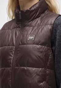 Близък план на човек, носещ тъмнокафяво подплътено елече с лого на GAP на гърдите и тъмна риза с дълъг ръкав под него.