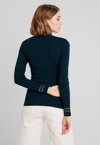 Donkergroene ribgebreide top met lange mouwen, een hoge kraag en subtiele metallic streepaccents op de manchetten. Gestructureerd materiaal en een nauwsluitend ontwerp.