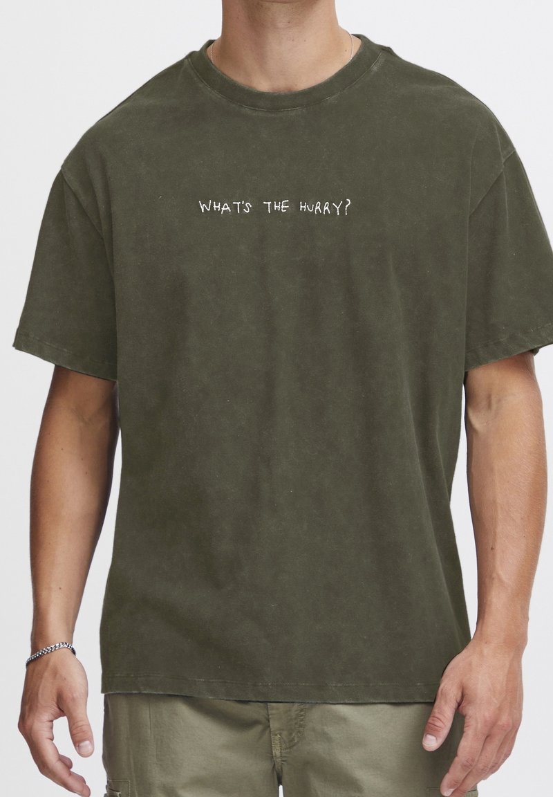 Homme portant un t-shirt vert olive avec texte blanc "WHAT'S THE HURRY?" et pantalon beige, montré du cou jusqu'à la mi-cuisse.