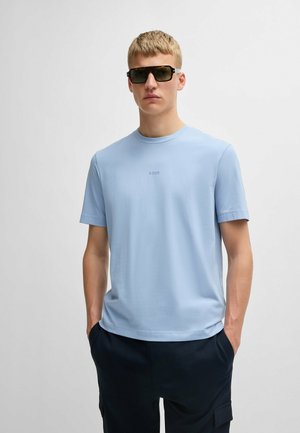 Helles blaues T-Shirt mit kurzen Ärmeln aus weichem Stoff, rundem Ausschnitt und dezentem Logodetail auf der Brust, kombiniert mit dunklen Hosen und Sonnenbrille.