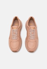 Ljusrosa lädersneakers med rund tå, silverfärgade snören och en guldprägling av "BOSS"-logotypen på tungan. Mjuk textur och minimalistisk design.