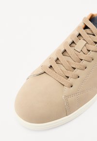 Beige leather sneaker with round toe, textured surface, matching laces, and white rubber sole. Features orange accent near the tongue.