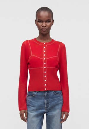 Kvinde iført en tætsiddende rød cardigan med knapper og hvide syningsdetaljer samt blå jeans, stående mod en ensfarvet lys baggrund.