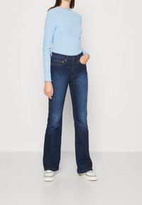 Hellblaues, geripptes Langarmshirt mit gewellten Bündchen, kombiniert mit dunkelblauen Schlagjeans und weißen Sneakers. Glatte Texturen und taillierte Passform.