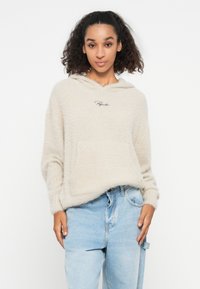 Beige, fluffy hoodie met een voorzak en geborduurde tekst. Draag het met lichtblauwe, losse jeans. Zachte textuur.