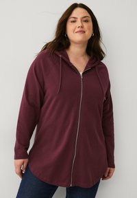 Burgunderfarbener Reißverschluss-Hoodie mit langen Ärmeln, Kordelzügen, weichem Stoff und abgerundetem Saumdesign. Kombiniert mit dunkelblauen Jeans.