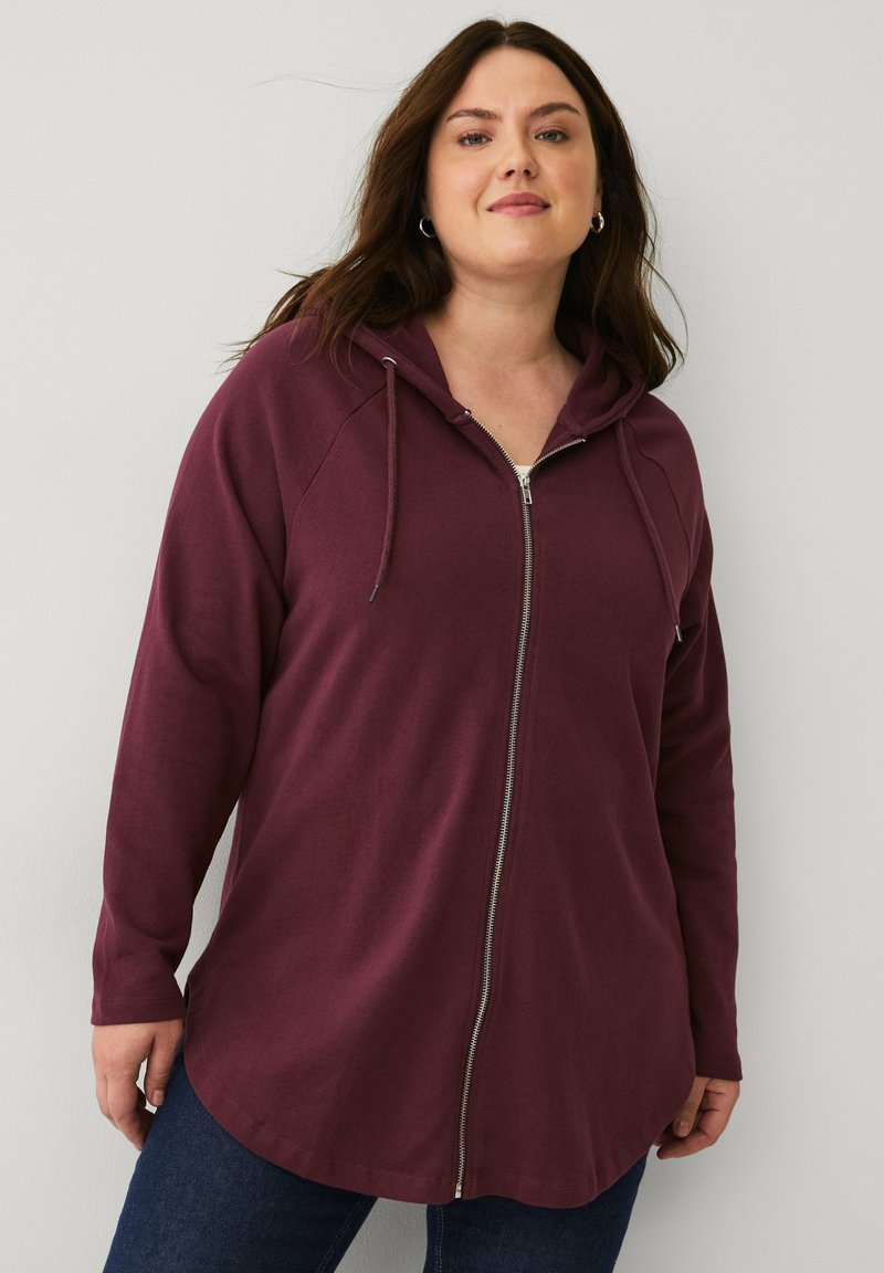 Burgunderfarbener Reißverschluss-Hoodie mit langen Ärmeln, Kordelzügen, weichem Stoff und abgerundetem Saumdesign. Kombiniert mit dunkelblauen Jeans.