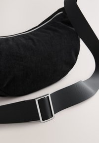 Zwarte corduroy tas met een zilveren rits en een brede, verstelbare zwarte riem met een rechthoekige metalen gesp. Gladde textuur.