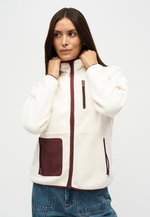 Une femme ajuste le col d’une veste polaire blanche ornée de fermetures éclair et de poches bordeaux, portée avec un jean bleu.