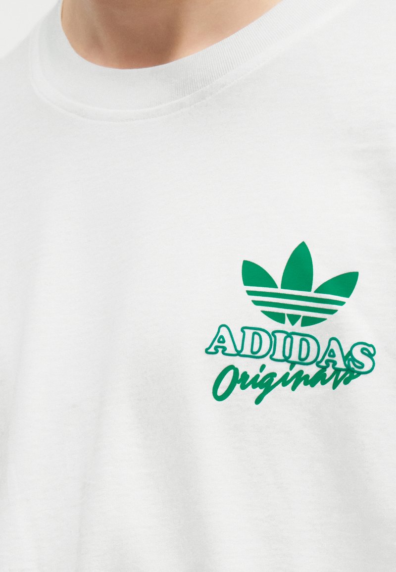 T-shirt blanc avec logo trèfle vert Adidas Originals et texte sur le côté gauche de la poitrine, porté par une personne.