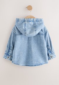 Hellblauer Denim-Kapuzenjacke für ein Kind, die an einem runden Holzwandhaken vor weißem Hintergrund hängt.