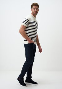 Sweter z krótkim rękawem w paski w granatowym i kremowym kolorze, zestawiony z ciemnymi spodniami w slim fit i czarnymi sneakersami, stojący na jednolitym tle.