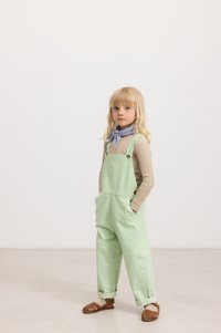 Grün gestreifte Latzhose mit Seitentaschen und verstellbaren Trägern, getragen über einem beigen Langarmshirt, ergänzt durch einen blauen Schal und braune Sandalen.